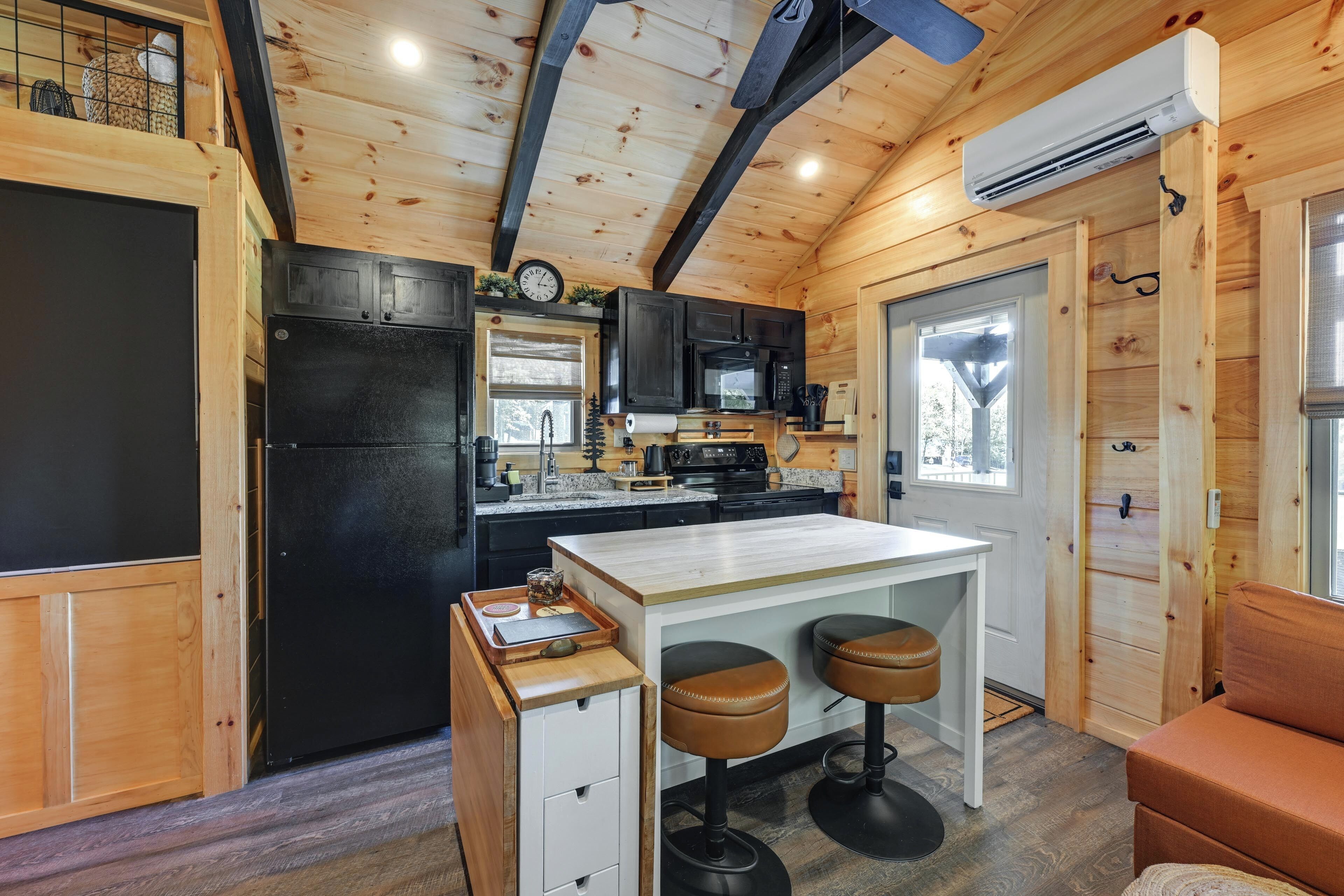 A compact kitchen with wooden cabinetry, a black refrigerator, a microwave, and a small island with stools.