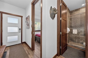 Ein Hotelzimmer mit Holztür, Duschkabine mit Glastür, Bett mit rot-schwarzem Karomuster und einem Fenster.