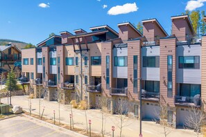 A row of modern multi-story residential buildings with balconies, a clear sky, and a parking area with no visible vehicles. - Arrow At Winter Park Unit 256 Downtown mt views, private HT, garage parking (Winter Park)