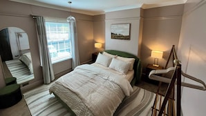 Ein Hotelzimmer mit Bett, Nachttischlampen, einem Fenster mit Vorhängen, einem Spiegel, einem Bild an der Wand und einem Sessel.
