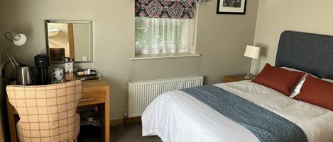 Habitación de hotel con cama, escritorio y silla, radiador, ventana con cortinas estampadas, un cuadro en la pared y televisor montado en la pared.
