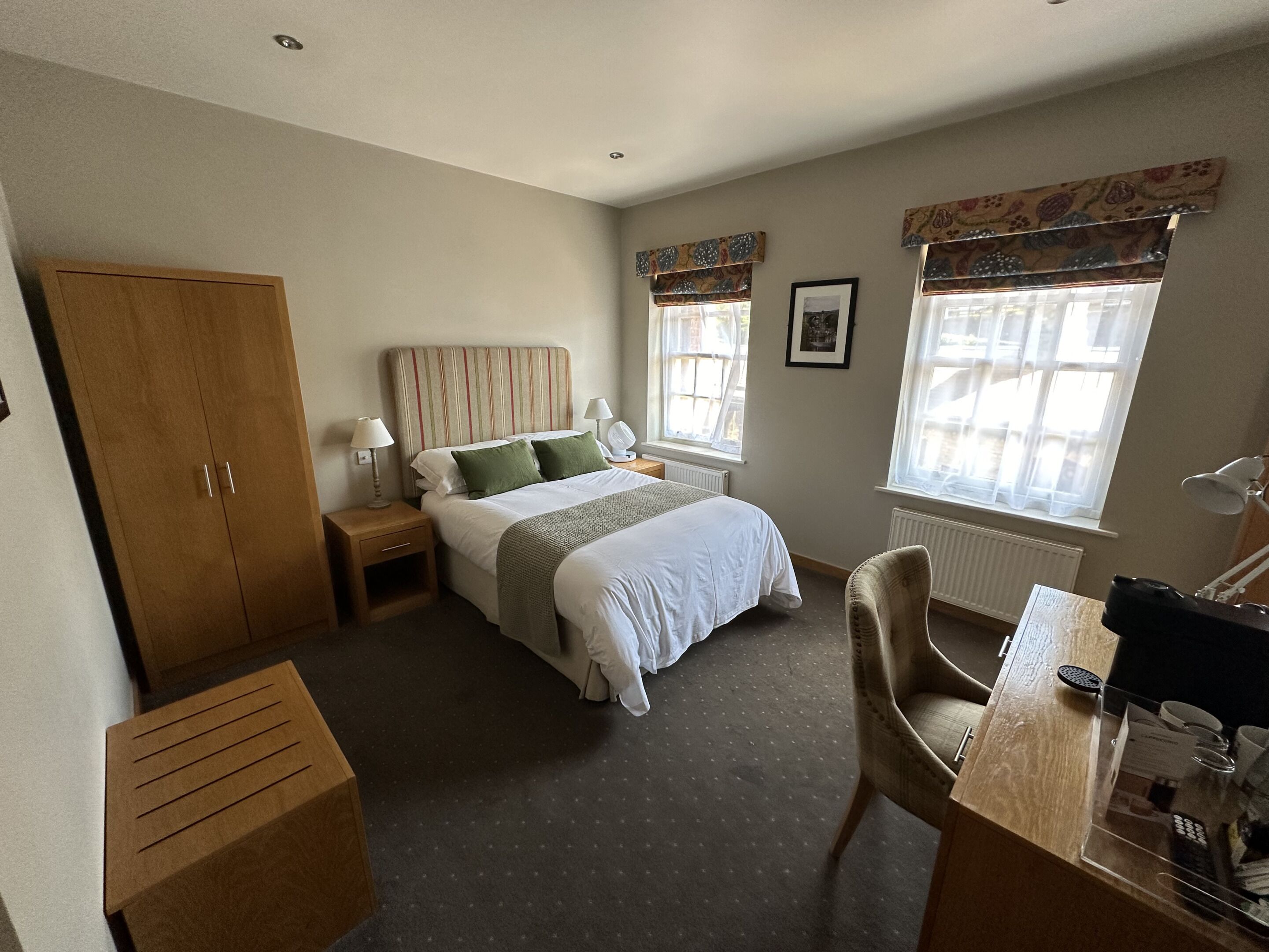 A bedroom with a bed, bedside tables, a wardrobe, a desk with a chair, and two windows with curtains.