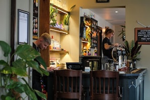 Uma área de bar com cadeiras de madeira, um balcão com um barman, prateleiras com garrafas e um letreiro na parede.