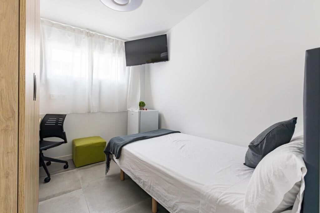 A small, modern bedroom with a bed, a desk, a chair, a TV, and a small refrigerator.