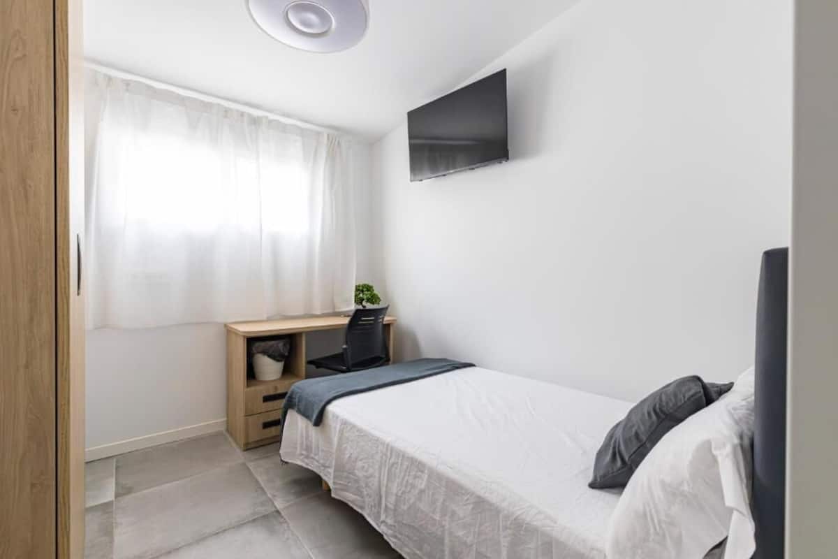 A bedroom with a bed, a desk, a chair, a TV, and a window with curtains.