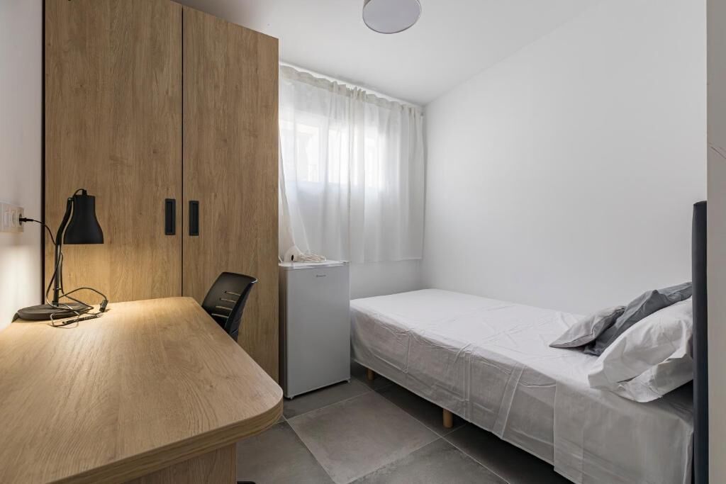 A small, modern room with a wooden wardrobe, a desk with a lamp, a bed, and a window with curtains.