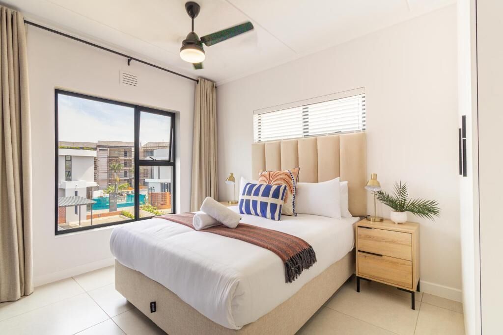 A modern hotel room with a large bed, a nightstand, a ceiling fan, a window with blinds, and a view of buildings and a pool.