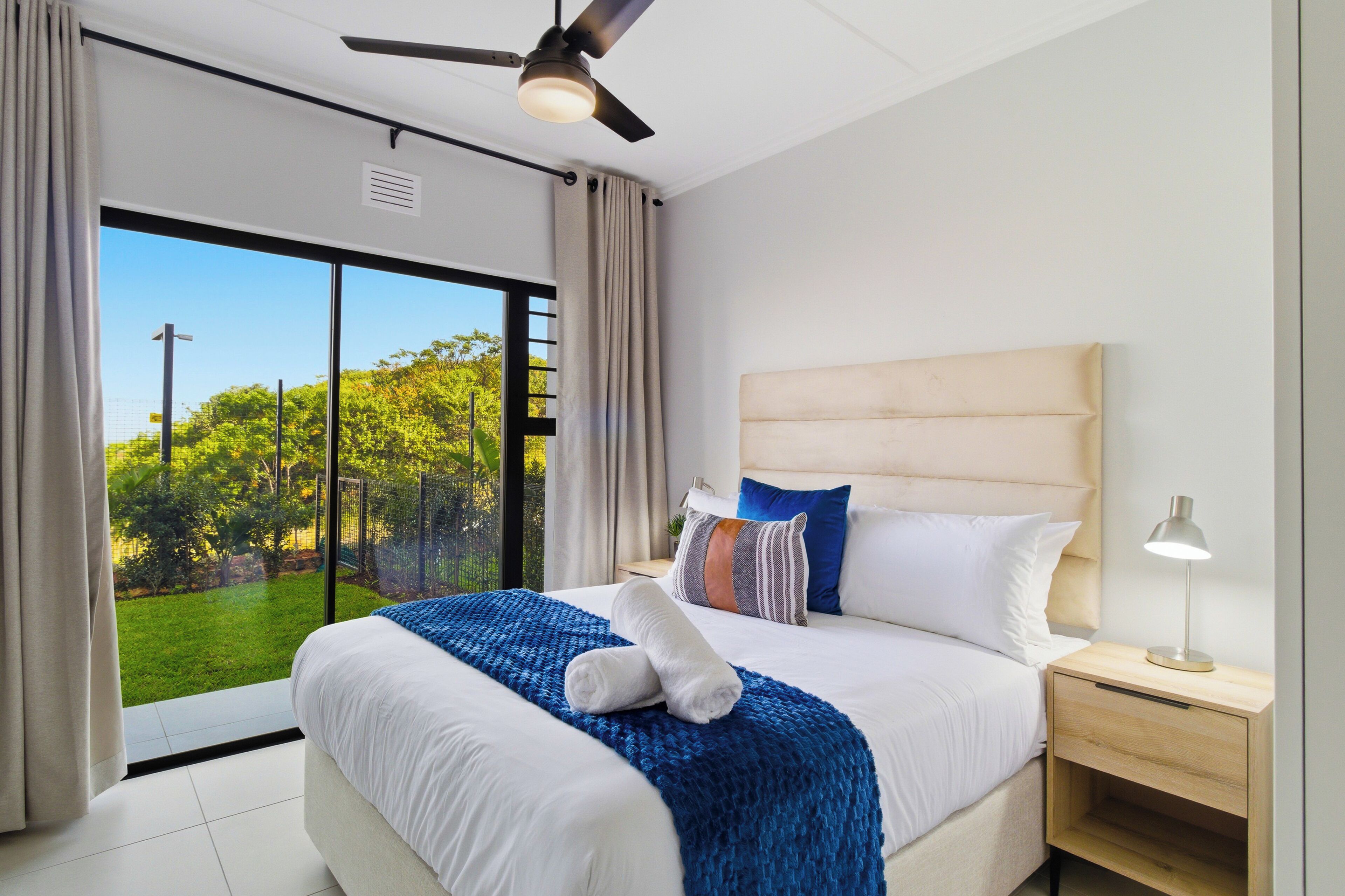 Habitación de hotel con cama, mesita de noche, ventilador de techo y un ventanal con vistas a la vegetación.