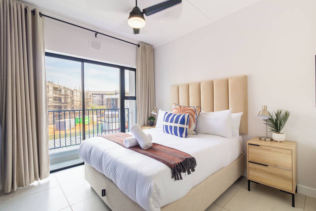 Un dormitorio con una cama grande, una mesita de noche con lámpara, una ventana con cortinas y un ventilador de techo.