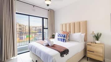 A modern bedroom with a large bed, a bedside table, a ceiling fan, and a view of the outside.
