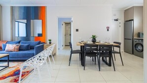 Uma sala de estar moderna com um sofá azul, um espaço de jantar com mesa e cadeiras de madeira, e uma cozinha com frigorífico e máquina de lavar.
