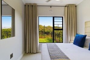 A hotel room with a large bed, a view of greenery through a sliding door, and a ceiling fan.