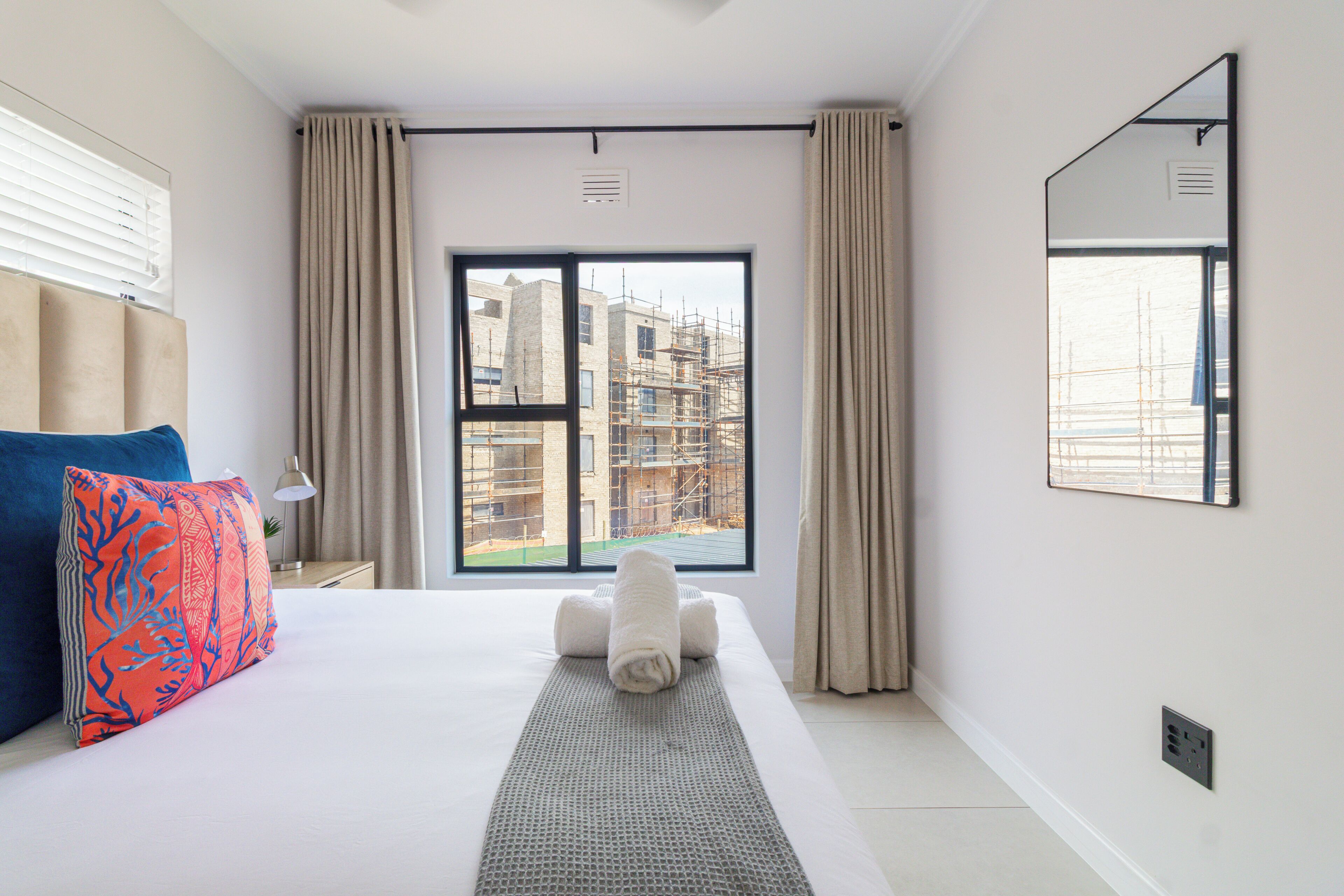 A hotel room with a large bed, a view of buildings through the window, a mirror on the wall, and a bedside lamp.