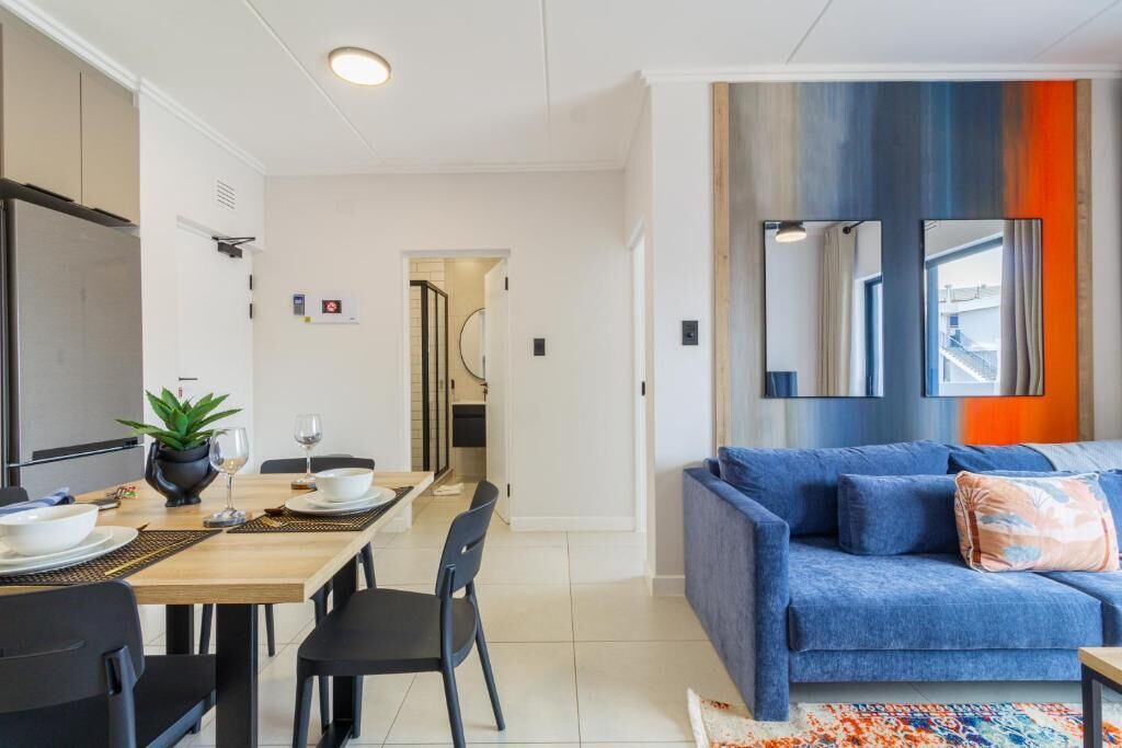 A modern living room with a blue sofa, a dining area with a table set for two, and a kitchen area with cabinets and a refrigerator.