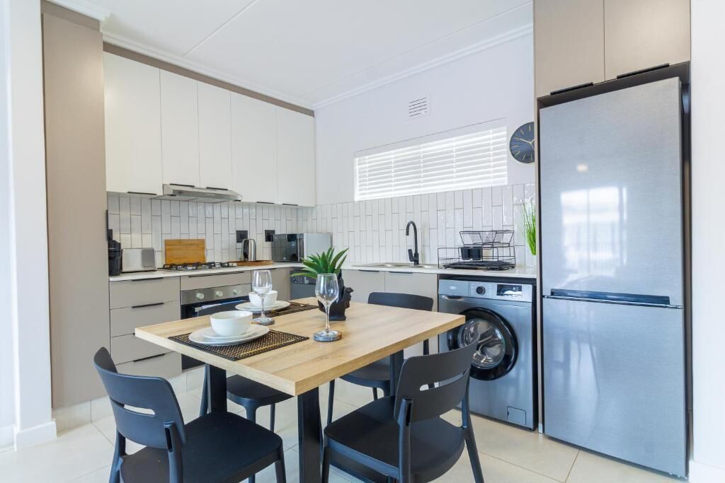 A modern kitchen with white cabinets, a stainless steel refrigerator, a washing machine, and a dining area with a table and chairs.