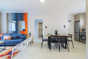 Una sala de estar moderna con un sofá azul, una mesa de comedor con sillas, una zona de cocina con refrigerador y lavadora.