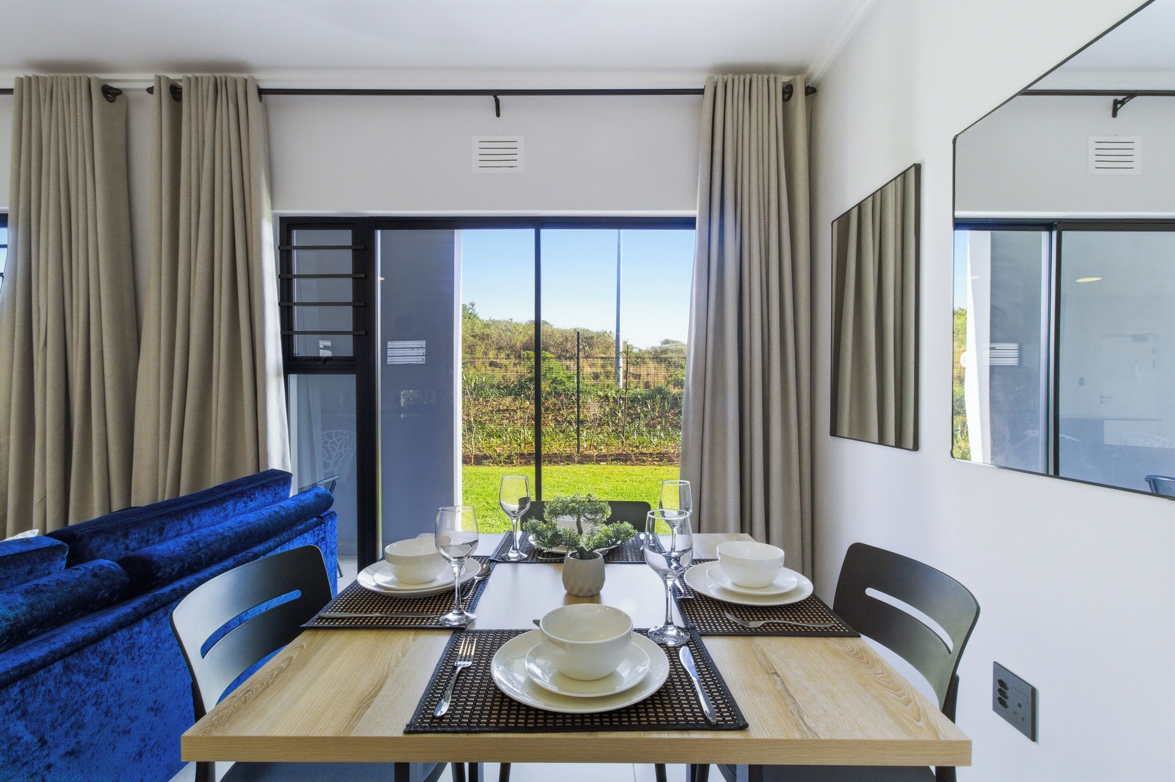 A dining area with a table set for four, a blue sofa, and a view of a garden through a large window.