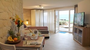 A hotel room with a bed, a TV, a dining area with a table and chairs, a small balcony with a view of the sea, and a stone wall. - Studio with Volcano View at Maarberg Resort in Schalkenmehren by the Lake (Schalkenmehren)