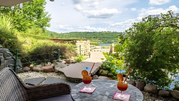 A patio with a wicker chair, two drinks on a table, a stone wall, and a view of a lake and hills. - Studio with Volcano View at Maarberg Resort in Schalkenmehren by the Lake (Schalkenmehren)
