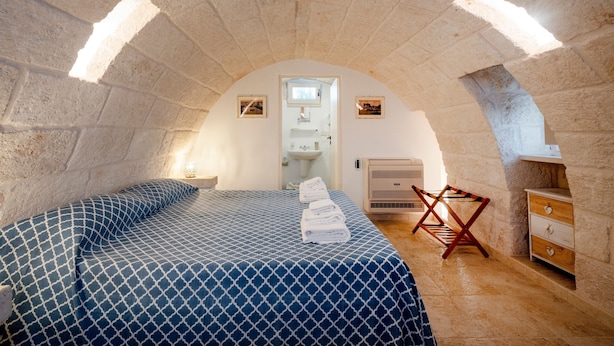 Ein Schlafzimmer mit Steindecke, einem Bett mit gemustertem Überwurf, einem Holzstuhl, einer Kommode und einem Waschbecken im Hintergrund.