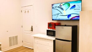 Una habitación compacta con una puerta blanca, una pequeña kitchenette con refrigerador, microondas y un electrodoméstico rojo, y un televisor de pantalla plana montado en la pared.