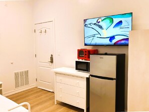 A compact room with a white door, a small kitchenette with a refrigerator, microwave, and a red appliance, and a flat-screen TV mounted on the wall.