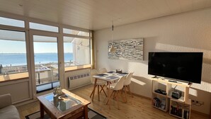 A modern living room with a large window overlooking a body of water, a dining area with a table and chairs, a flat-screen TV, and a bookshelf.