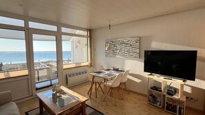 Ein modernes Wohnzimmer mit großem Fenster mit Blick auf ein Gewässer, einem Essbereich mit Tisch und Stühlen, einem Flachbildfernseher und einem Bücherregal.