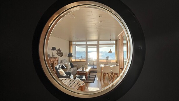 A round mirror reflecting a living room with a view of the sea, a dining area, and a sofa.