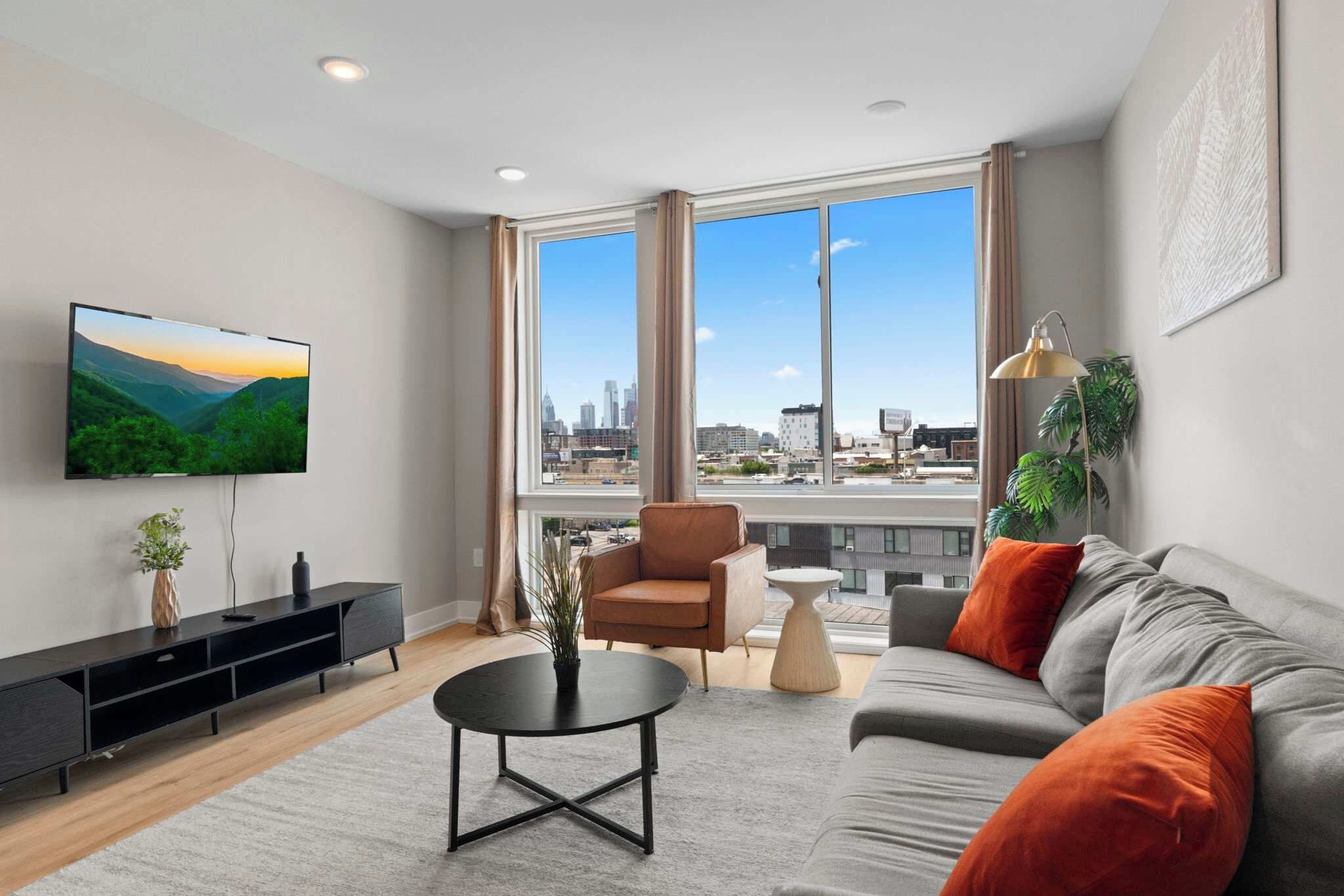 A modern living room with a large TV, a sofa, a coffee table, and a view of the cityscape through large windows.