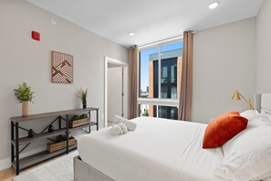 Een moderne hotelkamer met een groot bed, een console tafel met planten, uitzicht op de stad, en een raam met gordijnen.