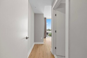 Un pasillo estrecho con suelo de madera y paredes blancas, que conduce a un balcón con vistas a edificios y un cielo despejado.
