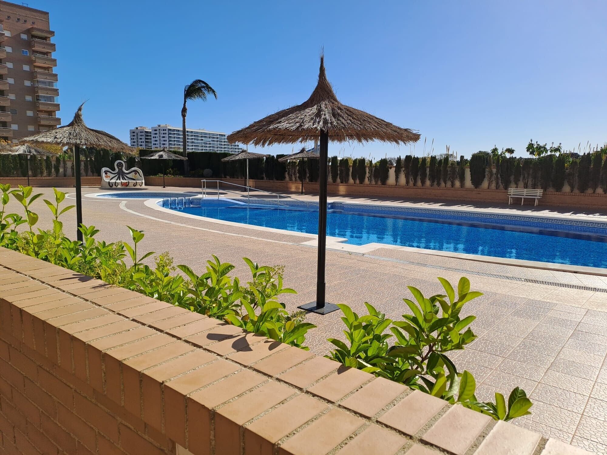 A swimming pool with a clear blue water, surrounded by a brick walkway, palm trees, and thatched umbrellas.. Apartment 'Frontal Jardines Del Mar 2' with Sea View, Wi-Fi and Air Conditioning