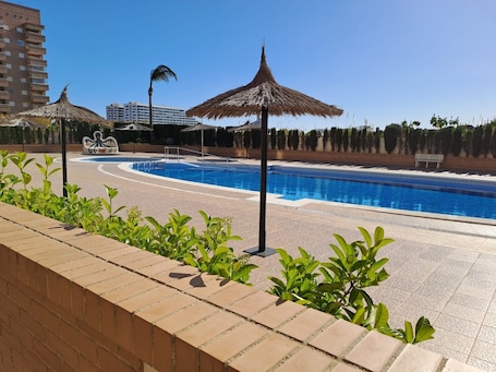 A swimming pool with a clear blue water, surrounded by a brick walkway, palm trees, and thatched umbrellas.. Apartment 'Frontal Jardines Del Mar 2' with Sea View, Wi-Fi and Air Conditioning