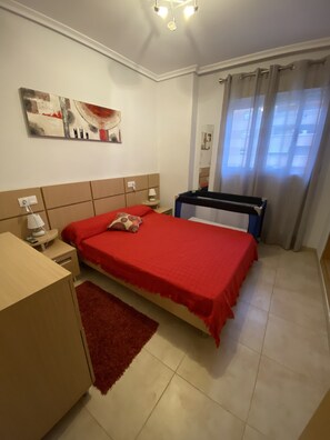 Ein Schlafzimmer mit einem Bett, einem Schreibtisch, einem Stuhl, einem Nachttisch, einer Lampe, einem roten Teppich, einem Gemälde und einem Fenster mit Vorhängen.