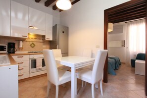 A compact kitchen with white cabinets, a refrigerator, a gas stove, and a coffee maker. There is a dining table with white chairs. - Puccini Historical Center (Venice)