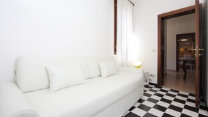 Un sofà blanc amb una làmpada, un paviment a escac i llambordes i una porta que dona a una altra estança.