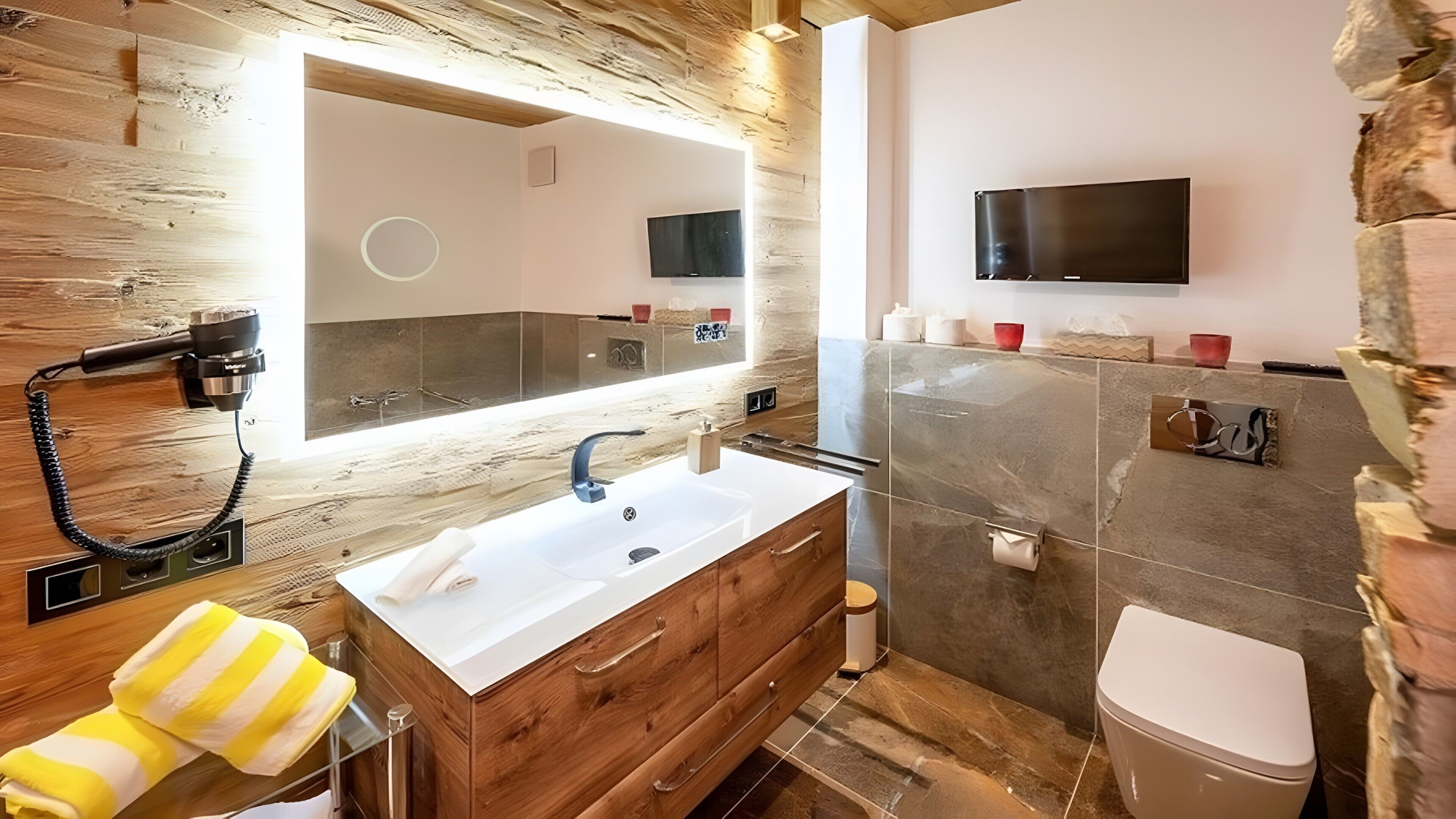Une salle de bain moderne avec un meuble-lavabo en bois, un grand miroir, un téléviseur fixé au mur et des toilettes.