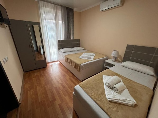 A hotel room with two beds, a wardrobe, a mirror, a window with curtains, and an air conditioning unit.