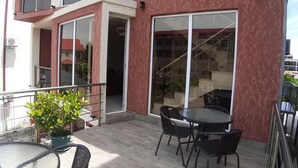 Un balcón con una mesa y sillas, una planta en maceta, y un edificio de fachada roja y ventanas amplias.
