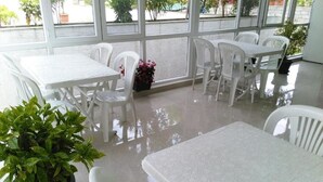 Un espacio interior limpio con mesas y sillas de plástico blanco, ventanas amplias y una planta en maceta.