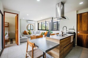 Una cuina moderna amb una illa central, una zona de menjador amb taula i cadires de fusta, un sofà integrat i un finestral gran que aporta llum natural.