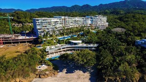 Et hotel med flere etager og poolområde, beliggende tæt på en strand med klart vand og et bjergrigt landskab.