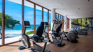 Et fitnesscenter med træningsmaskiner og store vinduer med udsigt over en strand.