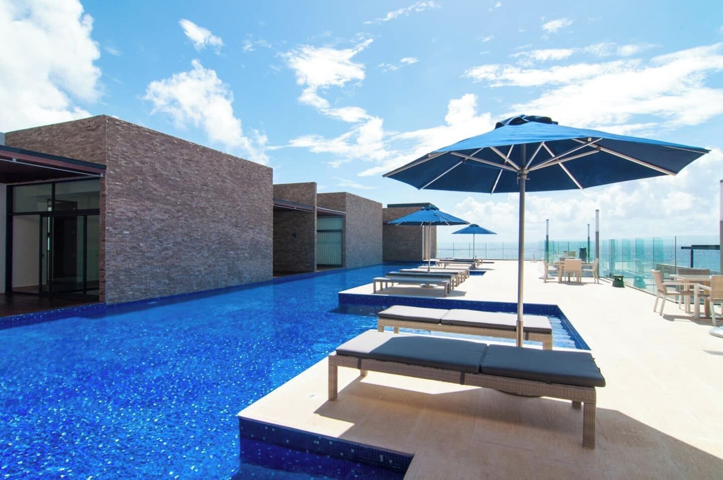 Una piscina infinita en la azotea con sillas de descanso y sombrillas, rodeada de arquitectura moderna.