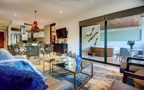 Ein modernes Wohnzimmer mit Sofa, Couchtisch und Sesseln, im Hintergrund eine Küche und ein Balkon mit Essbereich.
