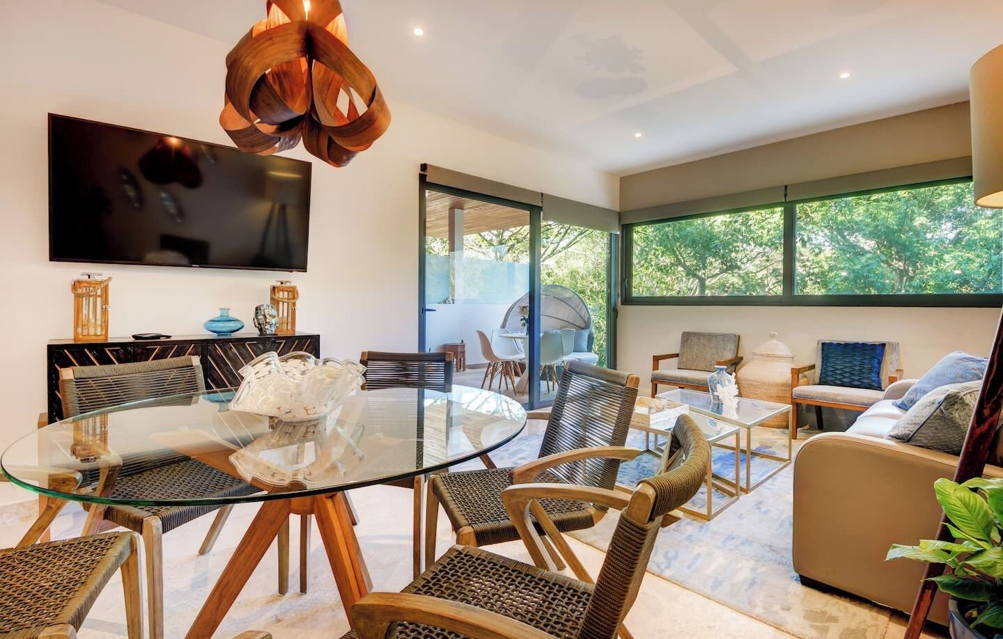 A modern living room with a glass dining table, wicker chairs, a sofa, and a TV mounted on the wall.