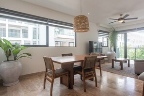 Ein Essbereich mit Holztisch und Stühlen, einer Hängeleuchte, einem Deckenventilator und großen Fenstern mit Blick auf Gebäude.