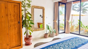 A bedroom with a wooden door, a large mirror, a bed with a patterned bedspread, and a view of an outdoor area with plants and a chair. - 2BR Private Jungle Pool Villa in Ubud · Boho Retreat, Yoga Corner & Lush Garden (Tampaksiring)