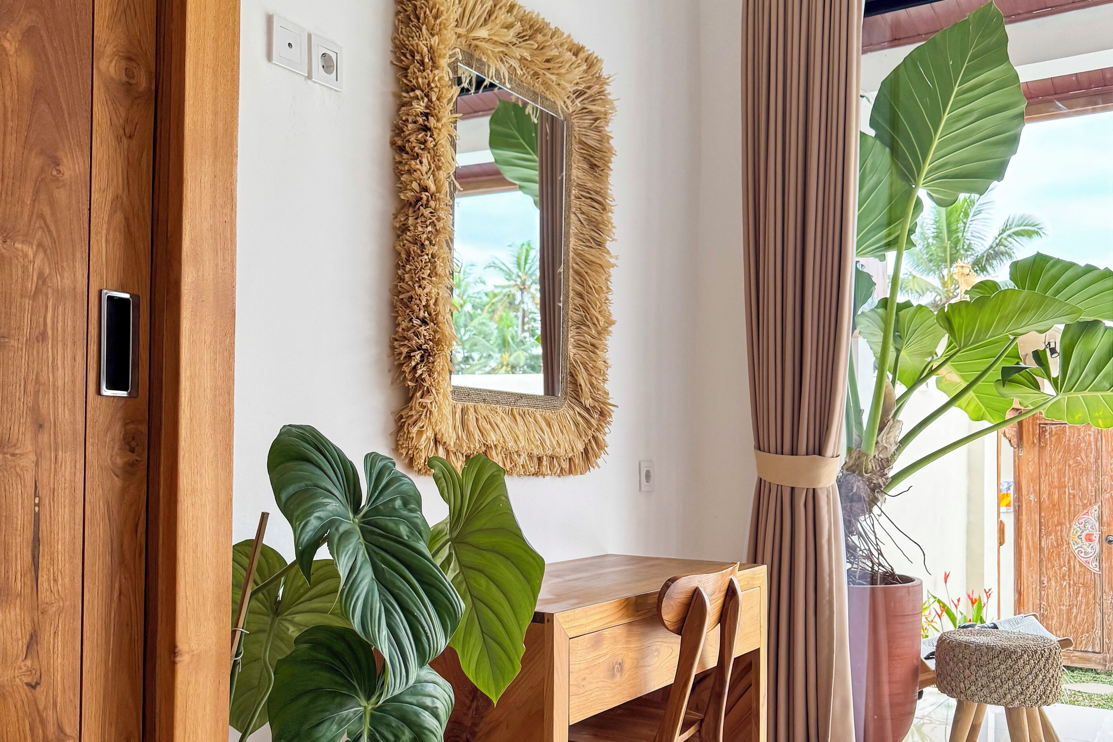 Een houten bureau met een spiegel, grote groene planten en een houten deur.